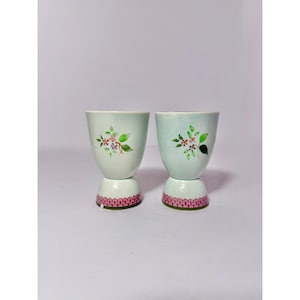 May include: Two light green ceramic egg cups with floral designs. The cups have a pink and white patterned base. The floral design includes pink and white flowers with green leaves. The egg cups are on a white background.
