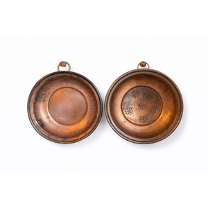 May include: Two copper bowls with a dark patina. One bowl is solid, and the other has small holes. Both have a brass ring and a leather loop for hanging. The bowls are round and have a slightly raised rim.