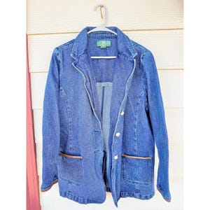 May include: A medium-wash blue denim blazer featuring a notched lapel, three front buttons, and two patch pockets with brown leather-like trim at the openings. The jacket is displayed on a white plastic hanger against a light-colored wall.