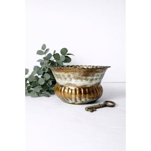 May include: A vintage-style metal bowl with a scalloped rim and a textured, aged appearance. The bowl has a gold and white color scheme, with a key resting nearby. Green foliage adds a natural accent to the composition.