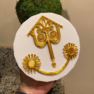 May include: A white round plaque with a gold-coloured design of a stylised house with two sun-like shapes on either side. The plaque is held in a hand.