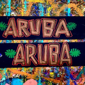K&ouml;nnte beinhalten: Zwei schwarze Holzpfeil-Schilder mit dem Wort "ARUBA" in braunen Buchstaben, die rot umrandet sind. Auf jedem Schild befinden sich gr&uuml;ne Blattmotive. Die Schilder befinden sich auf einem farbenfrohen, abstrakten Hintergrund.