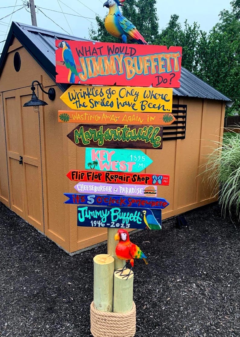 K&ouml;nnte beinhalten: Ein h&ouml;lzerner Wegweiser mit farbenfrohen Richtungsschildern, die S&auml;tze wie "Margaritaville" und "Flip Flop Repair Shop" enthalten. Der Wegweiser ist mit Papageienfiguren und dem Text "What Would Jimmy Buffett Do?" und "Jimmy Buffett 1946-2023" geschm&uuml;ckt.