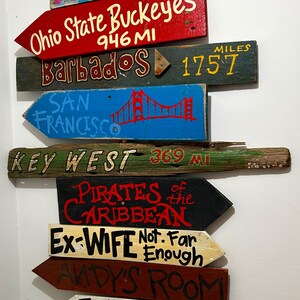 K&ouml;nnte beinhalten: Eine Sammlung von Holzpfeil-Schildern mit verschiedenen Zielen und Phrasen, die darauf gemalt sind. Zu den Zielen geh&ouml;ren "Pizza Planet", "Miami", "Ohio State Buckeyes", "Barbados", "San Francisco", "Key West", "Pirates of the Caribbean", "Ex-Wife", "Andy's Room", "Twilight Zone" und "Space Mountain".