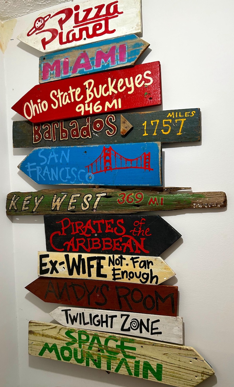 K&ouml;nnte beinhalten: Eine Sammlung von Holzpfeil-Schildern mit verschiedenen Zielen und Phrasen, die darauf gemalt sind. Zu den Zielen geh&ouml;ren "Pizza Planet", "Miami", "Ohio State Buckeyes", "Barbados", "San Francisco", "Key West", "Pirates of the Caribbean", "Ex-Wife", "Andy's Room", "Twilight Zone" und "Space Mountain".