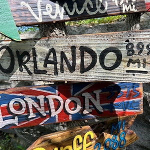 K&ouml;nnte beinhalten: Holzwegweiser mit aufgemalten Texten. Ein Schild zeigt nach Orlando, 1447 km entfernt. Ein anderes Schild zeigt die britische Flagge und weist nach London, 603 km entfernt. Weitere Schilder weisen nach Venedig und anderen Orten.