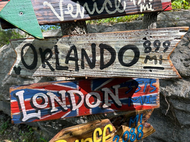 K&ouml;nnte beinhalten: Holzwegweiser mit aufgemalten Texten. Ein Schild zeigt nach Orlando, 1447 km entfernt. Ein anderes Schild zeigt die britische Flagge und weist nach London, 603 km entfernt. Weitere Schilder weisen nach Venedig und anderen Orten.