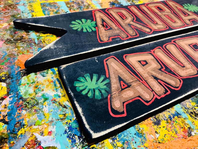 K&ouml;nnte beinhalten: Zwei schwarze Holzschilder mit dem Wort "ARUBA" in braunen Buchstaben, die rot umrandet sind. Jedes Schild hat ein gr&uuml;n gemaltes tropisches Blattdesign. Die Schilder befinden sich auf einem farbenfrohen, besprenkelten Hintergrund.