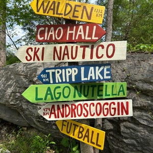 K&ouml;nnte beinhalten: Mehrere farbenfrohe Holzschilder, die an einem Baum befestigt sind und in verschiedene Richtungen weisen. Die Schilder tragen Namen wie "WALDEN", "CIAO HALL" und "SKI NAUTICO" sowie Entfernungen in Kilometern.