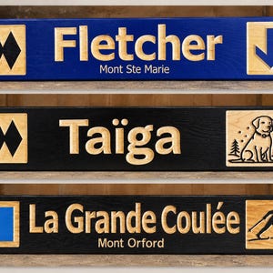 Peut inclure: Trois panneaux rectangulaires en bois avec du texte et des graphiques gravés. Le panneau supérieur est bleu avec "Fletcher Mont Ste Marie" et une flèche. Le milieu est noir avec "Taïga" et un chien. Le bas est noir avec "La Grande Coulée Mont Orford" et un skieur.