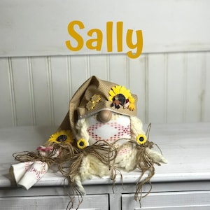 May include: A handmade gnome doll named "Sally" with a tan hat adorned with sunflowers. The gnome has a wooden nose, white yarn hair, and a patterned body. The doll is decorated with twine and small fabric accents.