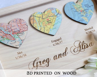 Può includere: Una targa in legno con tre mappe a forma di cuore, ciascuna rappresentante un luogo significativo nella relazione di una coppia. Le mappe sono di Vancouver, Staten Island e Manila. La targa include anche le date "Met 1/16/16", "Engaged 1/16/17" e "Married 9/18/18", insieme ai nomi "Greg e Alisa" e il testo "3D PRINTED ON WOOD".