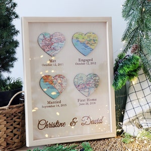 May include: A framed wooden sign with four heart-shaped maps representing milestones in a couple's relationship. The maps are labeled "Met", "Engaged", "Married", and "First Home" with corresponding dates. The names "Christine & David" are inscribed at the bottom.