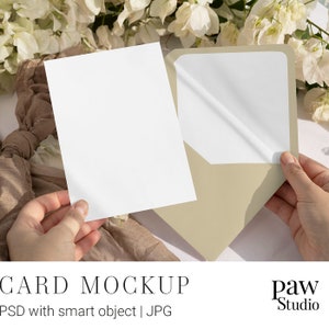 Puede incluir: Una maqueta de tarjeta blanca con un sobre beige. La tarjeta está siendo sostenida por una mano y el sobre está siendo abierto por otra mano. El fondo es una superficie blanca con una tela marrón y flores blancas.