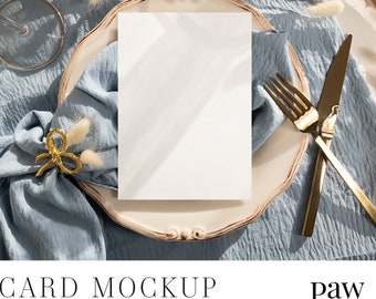 May include: A white card mockup on a blue and white plate with gold silverware. The plate is on a blue fabric background with white fluffy stems.
