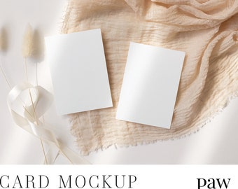 May include: Two blank white cards are arranged on a beige, textured fabric, with dried flowers and a cream-colored ribbon. The words "CARD MOCKUP" are at the bottom, along with "PSD with smart object | JPG" and the paw Studio logo.