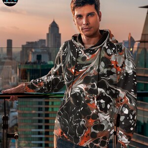 May include: A black hoodie with a swirling abstract pattern in shades of orange, white, and gray. The hoodie has a drawstring hood and a kangaroo pocket.