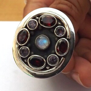 Puede incluir: Un anillo de plata con un gran engaste de forma ovalada. El engaste está lleno de diversas piedras preciosas, incluyendo una gran piedra de luna en el centro, rodeada de piedras más pequeñas de color rojo y morado.