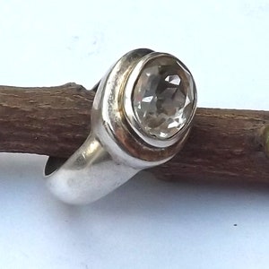 May include: A silver ring featuring a clear oval-shaped gemstone set in a bezel setting. The ring has a smooth, rounded band.