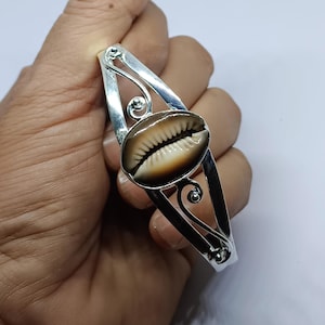 May include: A silver-toned cuff bracelet featuring a natural cowrie shell centerpiece. The shell is oval-shaped with a cream and brown pattern. The bracelet has decorative swirls and a polished finish, suitable for everyday wear.