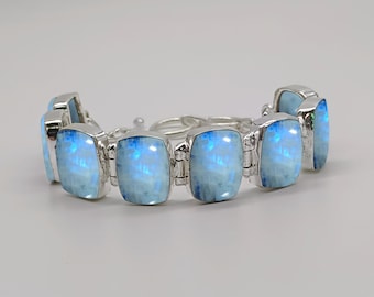 Könnte beinhalten: Ein Gliederarmband mit rechteckigen Mondstein-Cabochons in poliertem Silbermetall. Jeder Stein zeigt ein ausgeprägtes blaues Schimmern und ist durch silberne Scharniere verbunden. Das Armband enthält eine Silberkette zur Verlängerung und einen Karabinerverschluss.