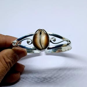 Op de afbeelding: Een zilverkleurige manchetarmband met een groot, ovaalvormig kaurieschelpmiddenstuk. De schelp is een mix van bruin en crème. De armband heeft een decoratief, open ontwerp met wervelende accenten aan elke kant.