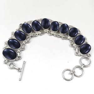 Op de afbeelding: Een zilveren armband met een kettingschakelontwerp en ovale blauwe stenen. De armband heeft een schuifgesp.