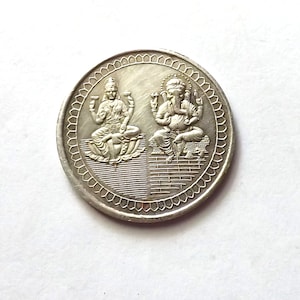 May include: A silver coin with an image of two Hindu deities, Lakshmi and Ganesha, on either side. The coin is surrounded by a decorative border.