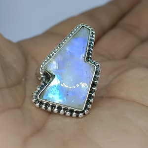 May include: A sterling silver ring featuring a large, irregular-shaped moonstone. The moonstone has a pearly white and blue iridescence. The ring's bezel is decorated with a beaded design and a twisted rope border.