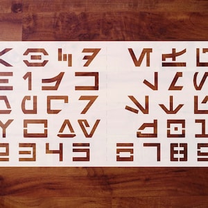 May include: White stencil with a set of 26 letters and numbers in a futuristic font. The letters are arranged in rows of four, with the numbers at the bottom.