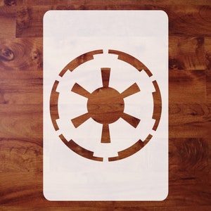 May include: A white stencil featuring the Galactic Empire symbol from Star Wars. The design is a circular emblem with a central sunburst pattern and a segmented outer ring. The stencil is rectangular with rounded corners.