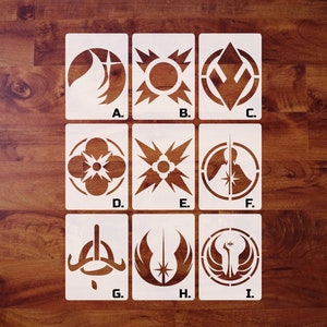 May include: Nine white stencils with various geometric and symbolic designs, labeled A through I, are displayed on a dark wood surface. The stencils feature shapes like circles, stars, and stylized figures, likely for crafting or artistic projects.