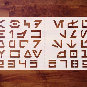 May include: White stencil with a set of 26 letters and numbers in a futuristic font. The letters are arranged in rows of four, with the numbers at the bottom.