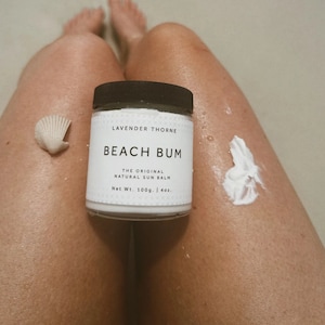 May include: A white jar of Beach Bum The Original Natural Sun Balm by Lavender Thorne. The jar is labeled with the brand name, product name, and net weight of 100g / 4oz.