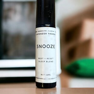 May include: A white roll-on bottle with a black cap and label. The label says "Lavender Thorne" and "Snooze Rest + Reset Sleep Blend" with a net volume of 10 ml.