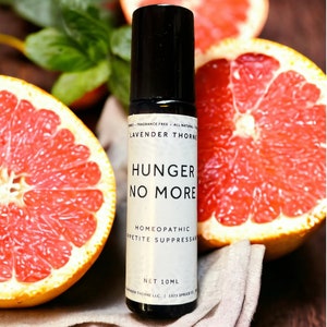 Puede incluir: Un frasco negro de roll-on con una etiqueta blanca que dice "LAVENDER THORNE HUNGER NO MORE HOMEOPATHIC APPETITE SUPPRESSANT NET 10ML". El frasco está sobre un paño blanco con dos mitades de pomelo.