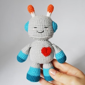 Puede incluir: Muñeca robot de ganchillo hecha a mano en tonos grises, azules, naranjas y rojos. El robot tiene una cara sonriente, un corazón rojo en el pecho y antenas. La muñeca mide aproximadamente 15 cm de altura.