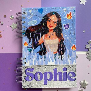May include: A blue and silver spiral notebook with a glittery cover. The cover features a cartoon illustration of a girl with long black hair, wearing a sparkly top and jeans. The notebook has the name "Sophie" written in purple letters on the front.