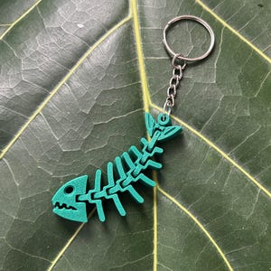 May include: A teal fish skeleton keychain with a silver chain and ring. The keychain is made of a solid material and is set against a dark green leaf.