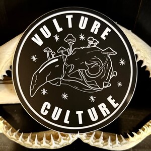 Peut inclure: Un autocollant rond noir avec les mots "VULTURE CULTURE" en blanc, entourant une illustration blanche d'un cr&acirc;ne avec des champignons. L'autocollant est orn&eacute; d'&eacute;toiles blanches.
