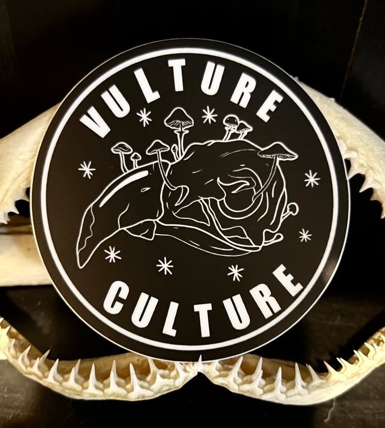 Peut inclure: Un autocollant rond noir avec les mots "VULTURE CULTURE" en blanc, entourant une illustration blanche d'un cr&acirc;ne avec des champignons. L'autocollant est orn&eacute; d'&eacute;toiles blanches.