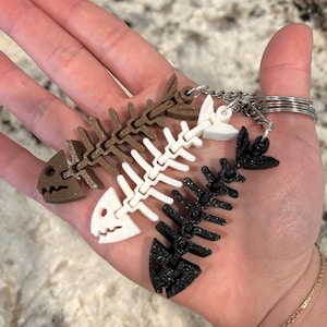 May include: Four fishbone-shaped keychains in brown, white, and black. Each keychain features a detailed fish skeleton design. The keychains are attached to a silver keyring.