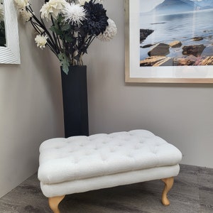 May include: A white tufted ottoman with wooden legs. The ottoman is sitting on a wooden floor.