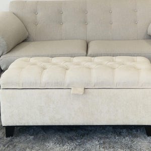 May include: A beige upholstered ottoman with a tufted top and a lift-up lid, positioned in front of a matching beige sofa. The ottoman has black legs and a rectangular shape, offering storage and seating.