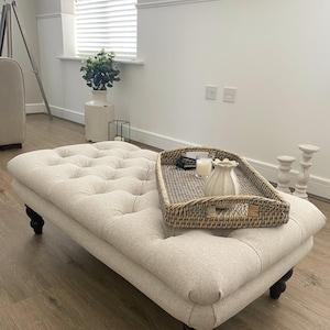 May include: A white upholstered ottoman with button tufting and dark wood legs. A woven wicker tray with a white ceramic vase and candles sits on top of the ottoman.
