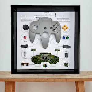 May include: Framed artwork showing a disassembled Nintendo 64 controller. The grey controller is presented alongside its internal parts, such as the circuit board, buttons, and joystick. The background includes technical details and release dates.