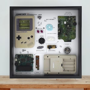 May include: A black shadow box frame containing a disassembled beige Game Boy console, including the front shell with a Tetris screen, the back battery compartment, green circuit boards, buttons, a speaker, and labels identifying each component.