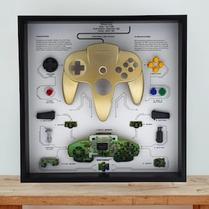 May include: A framed display featuring a disassembled gold Nintendo 64 controller and its internal circuit board. The components are arranged on a white background with technical text labels, including 'CONTROL PAD', 'START BUTTON', 'A AND B BUTTONS', and 'LOGIC BOARD'.