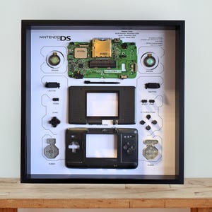 May include: A framed display showcasing a disassembled Nintendo DS console. The black frame encloses a white background with the console's components, such as the circuit board, buttons, and screens, meticulously arranged. The text "Nintendo DS" is displayed.