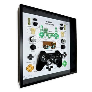 Pu&ograve; includere: Opera d'arte incorniciata con un controller Sony Dualshock 2 scomposto. Il controller nero &egrave; esposto con i suoi componenti, inclusi pulsanti, circuiti stampati e motori di vibrazione, su uno sfondo bianco. La cornice &egrave; nera.